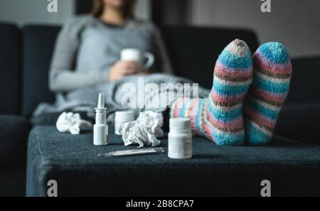 Sick woman resting on couch holding hot cup of tea. Ill person with flu, cold, fever or virus sitting on sofa at home in winter. Medicine, thermometer. Stock Photo