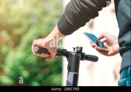 Electric scooter rent with mobile phone. Man using smartphone rental app in city street or park in summer. E kick vehicle. Modern transportation. Stock Photo