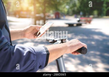 Electric scooter in city. Man using smartphone to rent an e kick vehicle. Millennial guy with mobile phone using eco transport in downtown. Stock Photo
