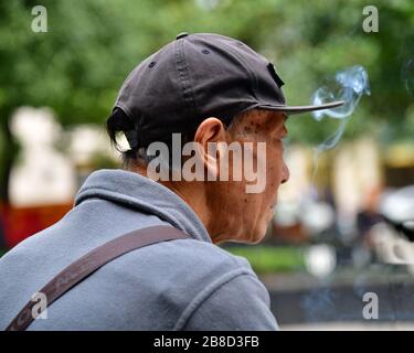 En profile photo of an old Chinese man sitting in a park in Shanghai is staring in the distance while smoking a cigarette and listening to music Stock Photo