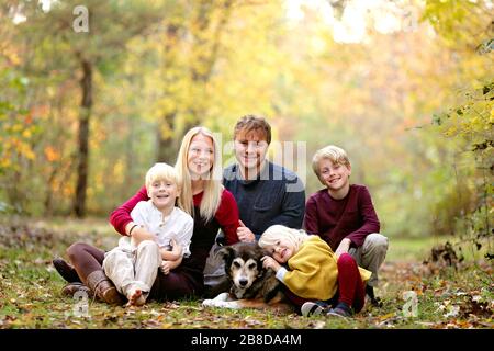 A portrait of a happy family of 5 people, inclluding three cute smiling children, sitting outside in the woods with their pet dog on an Autumn day. Stock Photo