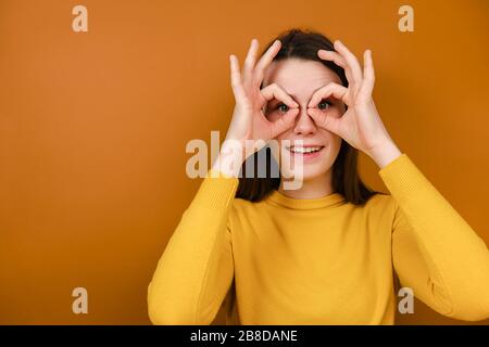 Funny smiling young woman having fun making glasses shape with hands, does funny face with eyes wide open, dressed in yellow sweater Stock Photo