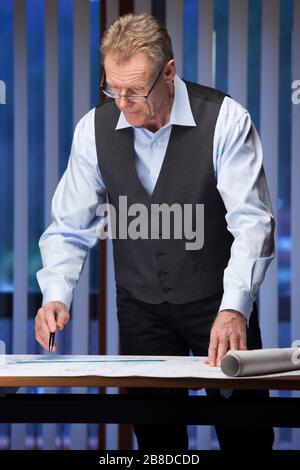 Mature engineer, architect or businessman standing at a desk in an office late at night looking at a plan Stock Photo