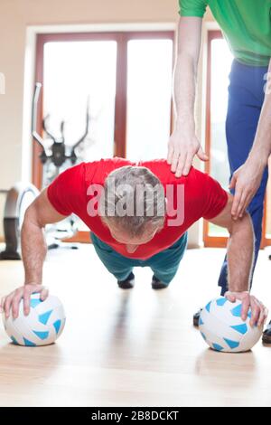 Mature man in red t-shirt doing push-ups  in a fitness center with the assistance of an instructor Stock Photo