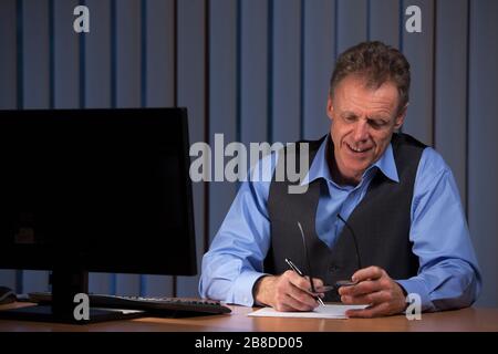 Mature businessman taking notes sitting at a desk with computer screen and smiling Stock Photo