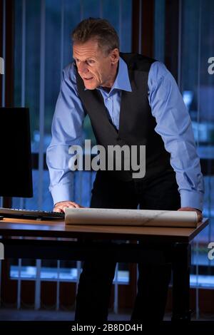 Senior architect or engineer standing at his desk looking at computer screen in a dark office at night Stock Photo