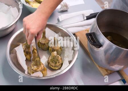 Subtract oil from caramelized artichokes on a bowl of absorbent paper towels. Woman hands with kitchen tongs. Stock Photo