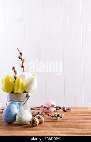 Congratulation festive composition from freshly picked spring colored tulips flowers in a bucket and handmade painted eggs on a wooden table against l Stock Photo
