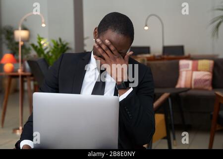 Total disappointment. Young African man looking exhausted and covering his face with hands while sitting at his working place Stock Photo