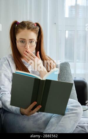 A redhead girl with glasses, with long tails in pajamas sits on the couch and looks in shock at the book, covering mouth with hand Stock Photo