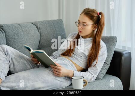 A red-haired girl with glasses, with long tails in pajamas lies on the sofa and carefully reads a book Stock Photo
