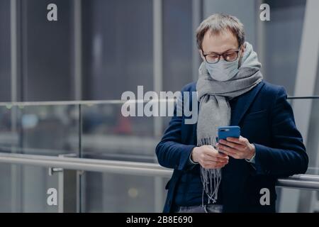 Horizontal shot of man in spectacles uses modern mobile phone, wears medical mask during coronavirus outbreak, has influenza protection in big shoppin Stock Photo