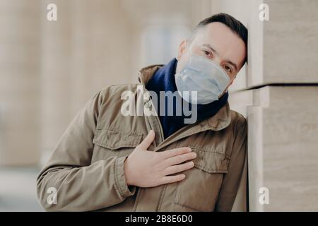 Sad young man wears protective medical mask, walks through city streets, protects himself from pandemic coronavirus, has health problems, hard breathi Stock Photo