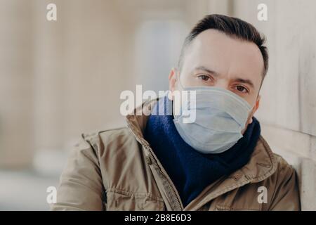 Outdoor shot of young European man has coronavirus symptoms, wears protective surgery mask, tries protect from viruses and infections, needs to see do Stock Photo