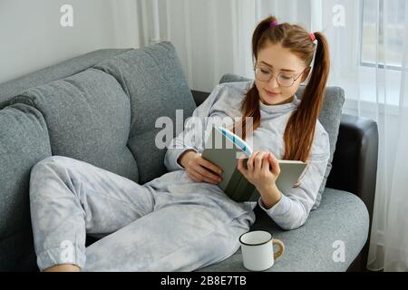 A red-haired girl with glasses, with long tails in pajamas lies on the sofa with a mug and reads a book Stock Photo