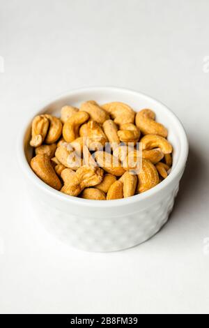 A small white bowl of delicious healthy salted cashew nuts isolated on a white background close up