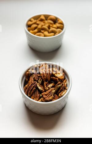 A small white bowl of delicious healthy salted pecan nuts in front of a white bowl of cashews isolated on a white background Stock Photo