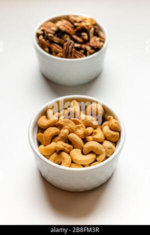 A small white bowl of delicious healthy salted cashew nuts in front of a white bowl of pecans isolated on a white background Stock Photo