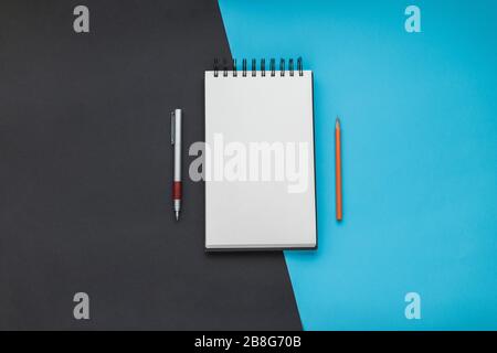 Open spiral sketchbook in the middle with a blank sheet, a pen and an orange pencil with a black and blue diagonal backdrop. Still life, office suppli Stock Photo