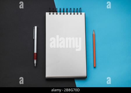 Open spiral sketchbook in the middle with a blank sheet, a pen and an orange pencil with a black and blue diagonal backdrop. Still life, office suppli Stock Photo