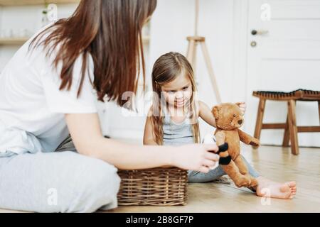 Cute toddler girl long fair hair big grey eyes wearing pajamas playing bear toy with mother at the room stay at home Stock Photo