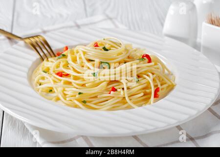 Pasta Aglio, Olio e Peperoncino, italian spaghetti with garlic, chili pepper and olive oil on a white plate with golden fork and salt shaker on a wood Stock Photo