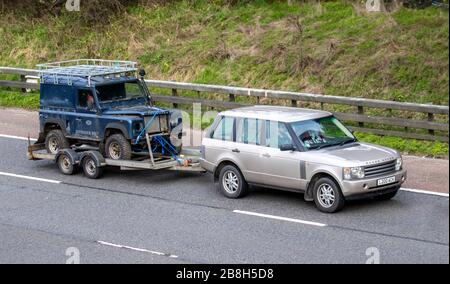 Range Rover towing decrepit dilapidated old Land Rover Defender. A restoration project, towed barn find; UK vehicular traffic, transport, moving vehicles, restoration vehicle, roads, towing motors, motoring trailer on the M6 motorway highway Stock Photo