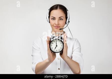 Beautiful smiling woman consultant of call center in headphones holding alarm clock on gray background. female customer support operator with headset. save your time Stock Photo