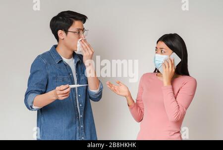 Coronavirus Anxiety. Asian Girl In Medical Mask Calling Ambulance For Sick Boyfriend Stock Photo