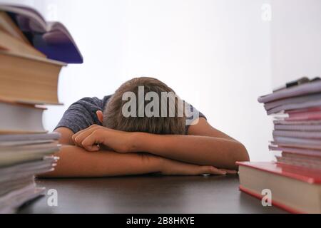 Tired disappointed schoolboy lowering his head sitting among pile of books, textbooks, school exercise books on his desk at home Stock Photo