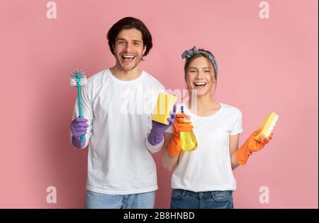 Excited couple wearing gloves, holding cleaning supplies in hands, pink background Stock Photo