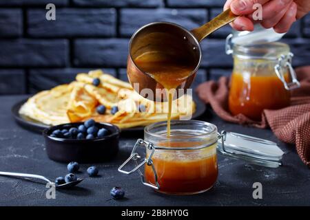 woman is pouring freshly cooked homemade buttermilk syrup from copper saucepan to a glass jar on a concrete table. crepes and a brick wall at the back Stock Photo