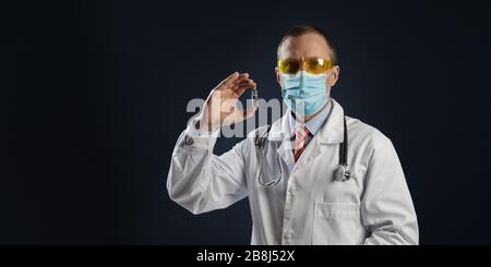 Mature male doctor holding a vaccine in his hands. The doctor in a medical protective mask and goggles. Stock Photo
