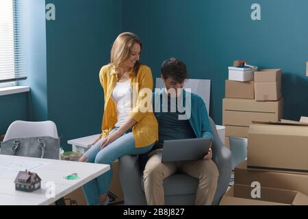 Happy young couple moving in their new home, they are sitting on the armchair together and connecting with their laptop, home relocation concept Stock Photo