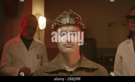 Scientists making experiments on a human brain in a vintage style lab, a man is wearing a sci-fi helmet and researchers are testing him Stock Photo