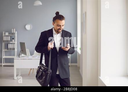 Young hipster businessman in black suit reads message in mobile standing in office. Stock Photo