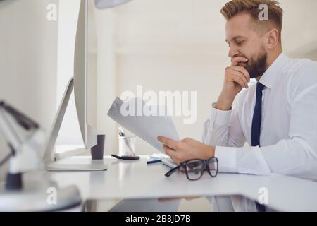 Serious busy businessman reads working documents sitting at a table with a computer in the office. Stock Photo