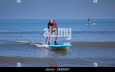 Portobello, Edinburgh, Scotland, UK. 22nd Mar 2020. Sunshine and blue skies with an early morning temperature of 2 degrees centigrade rising to 8 degrees by noon. Two stand up paddle boarders along with a variety of people out on the beach relaxing and enjoying activities to get away from the television and radio. Stock Photo