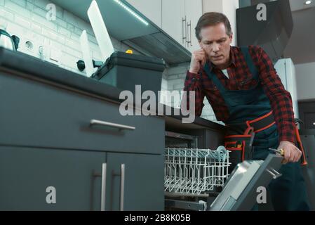 Portrait of mature man wearing tool kit standing in the kitchen, holding a screwdriver and thinking how to fix the dishwasher door. Horizontal shot Stock Photo