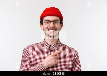 Close-up portrait handsome friendly-looking hipster male in red beanie and glasses, smiling satisfied and happy, pointing finger upper right corner Stock Photo