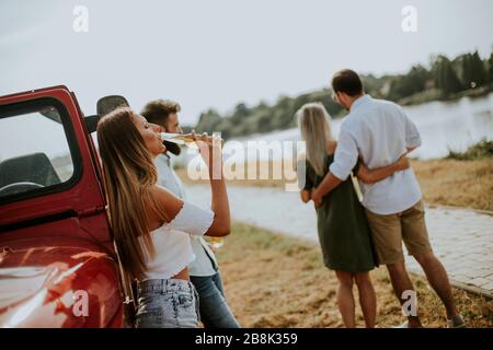 Happy young women drinks cider from the bottle by the convertible car with friends Stock Photo