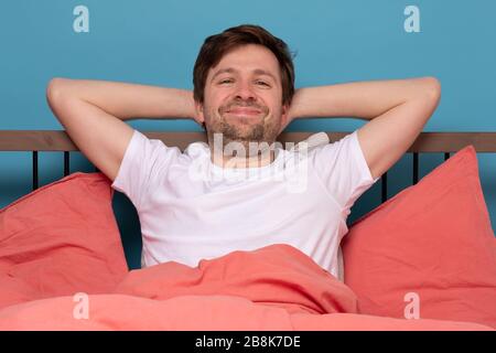 Young man holding smiling confident with crossed arms resting at bed alone at quarantine. Studio shot on blue wall. Stock Photo