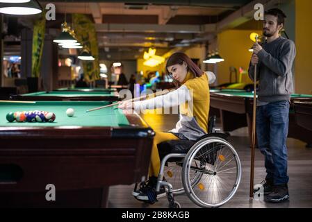 Beautiful disabled girl in a wheelchair playing billiards with her friend Stock Photo