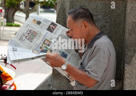 Close up of local man in Singapore out on the street, keeping in the shade, and reading a newspaper Stock Photo