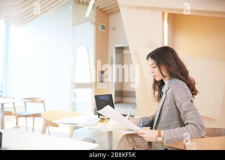 Warm-toned side view portrait of pretty young woman working or studying sitting at table in empty cafe hall, copy space Stock Photo