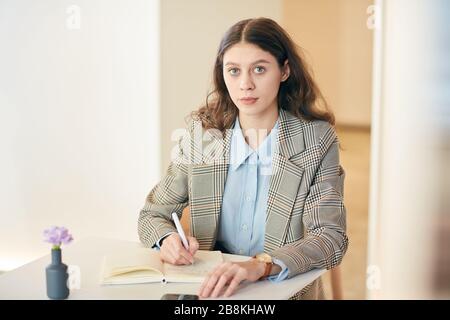 Portrait of pretty young woman looking at camera while taking notes sitting at white cafe table, copy space Stock Photo