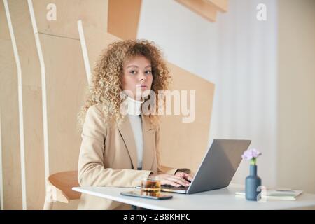 Portrait of modern curly haired young woman looking at camera while using laptop sitting at table in cafe, copy space Stock Photo