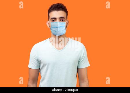 Portrait of serious handsome man with medical mask in casual white t-shirt standing with hands down and looking focused and confident at camera. indoo Stock Photo