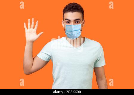 Hello! Portrait of friendly glad handsome man with medical mask in casual white t-shirt showing hi gesture with waving hand and smiling sincerely. ind Stock Photo