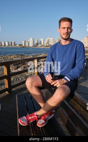 a handsome man drinking Yerba Mate Infusion in the rambla of Punta del Este, Maldonado, Uruguay with the atlantic ocean and city at the background Stock Photo